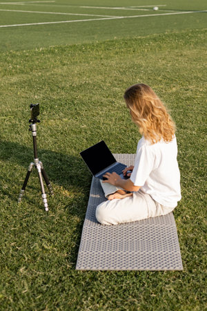 long haired yoga teacher using laptop while sitting near tripod with cellphone on grassy stadiumの写真素材