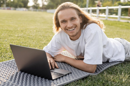 young and carefree man looking at camera while lying near laptop on yoga matの写真素材
