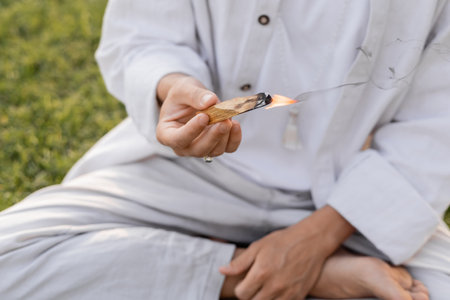 partial view of man in white cotton clothes holding burning palo santo stick while meditating outdoorsの写真素材