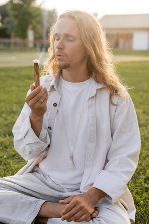 long haired man in white linen clothes blowing at aromatic palo santo stick during esoteric ritual outdoorsの写真素材