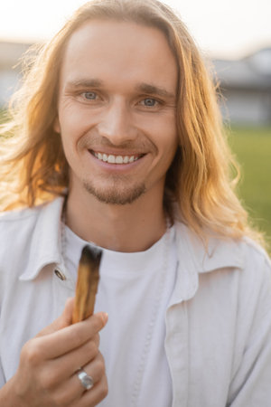 joyful long haired yoga man holding smoldering palo santo stick and smiling at camera outdoorsの写真素材