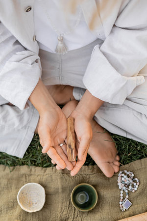 top view of cropped man in white clothes holding palo santo stick near linen rug with ceramic cups and mala beadsの写真素材