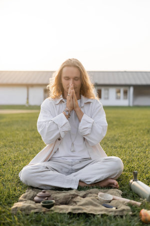 young man in white clothes meditating in easy pose near linen rug with clay teapot and bowls on green lawnの写真素材