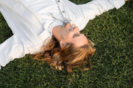 top view of carefree long haired man in white shirt lying and smiling on green lawnの写真素材