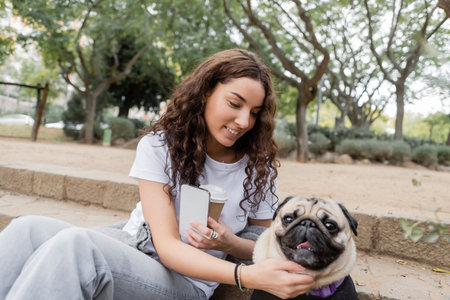 Pretty and curly young woman in casual clothes holding smartphone and coffee to go while petting pug dog on stairs and spending time in blurred park in Barcelona, Spainの写真素材
