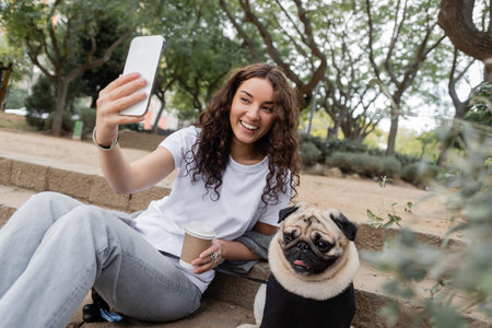 Carefree young and curly woman in casual clothes having video call on cellphone and holding coffee to go while sitting near pug dog on stairs in blurred park at daytime in Barcelona, Spainの写真素材