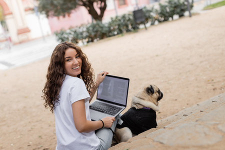 Cheerful young curly woman in casual clothes looking at camera while holding laptop near pug dog on...