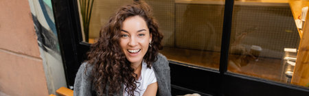 Smiling young and curly woman in casual jacket looking at camera while sitting near outdoor cafe on...