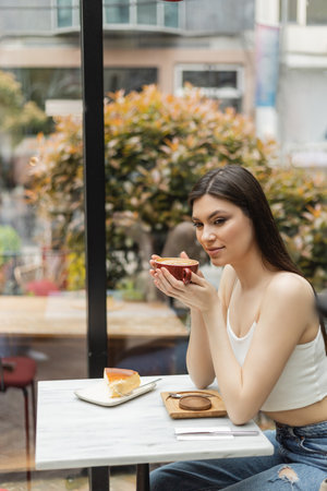 happy woman with long hair sitting next to window and holding cup of cappuccino with coffee art near cheesecake on plate of bistro table inside of modern cafe in Istanbulの写真素材