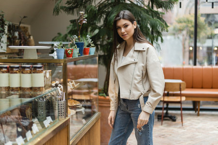 brunette young woman with long hair in beige leather jacket and denim jeans looking at camera while standing near cake display with pastry and jars of jam in modern bakery shop in Istanbulの写真素材