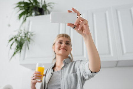 low angle view of happy woman with bangs and rings on fingers holding glass of orange juice and taking selfie on smartphone and standing in blurred white kitchen with green indoor plantsの写真素材