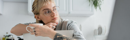 dreamy young woman with tattoo on hand and bangs holding cup of coffee and looking away near blurred laptop in modern kitchen, freelancer, remote lifestyle, eyeglasses, bannerの写真素材
