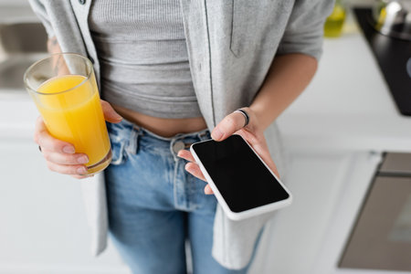 cropped view of young woman with tattoo on hand holding glass of fresh orange juice and smartphone with blank screen while standing in casual clothes with blue denim jeans in modern apartmentの写真素材