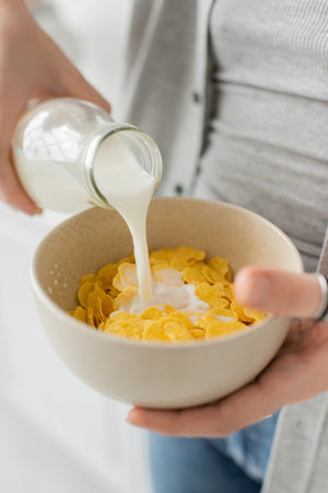 close up view of young woman holding bottle while pouring fresh milk into bowl with cornflakes and...