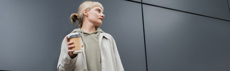 fashionable young woman with blonde hair with bangs standing in coat and hoodie while holding paper...
