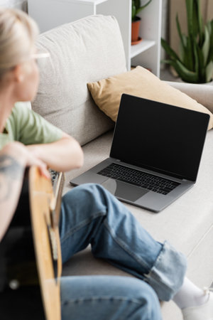 young woman in glasses holding acoustic guitar and learning how to play while looking video tutorial on laptop with blank screen and sitting on comfortable couch in living room, guitar lessonsの写真素材