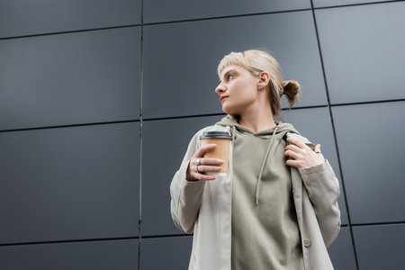 stylish young woman with blonde hair with bangs looking away and standing in coat and hoodie while holding paper cup with takeaway coffee near grey modern building on street, outside, urban livingの写真素材