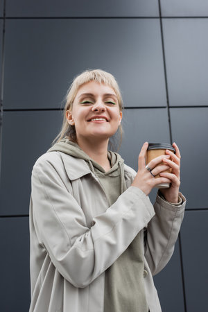 low angle view of happy young woman with blonde hair with bangs standing in coat and hoodie while...