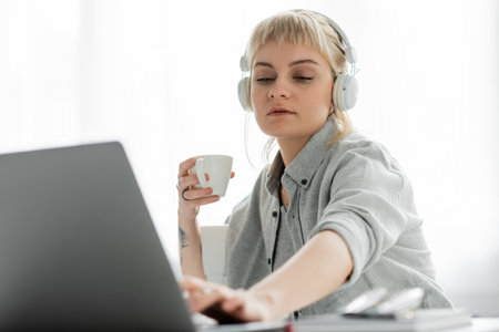 young freelancer with blonde hair, bangs and tattoo on hand sitting in wireless headphones and...