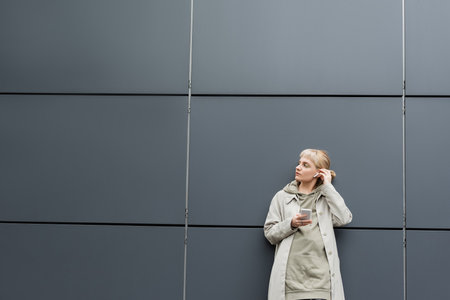 fashionable young woman with bangs standing in trendy clothes and wireless earphones while using smartphone near grey modern building on urban street, coat with hoodie, looking awayの写真素材