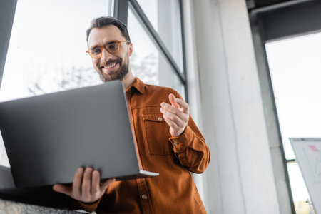 low angle view of pleased bearded entrepreneur in fashionable eyeglasses and shirt gesturing during...