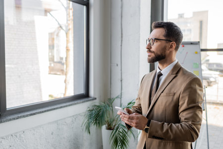 serious bearded businessman in beige blazer and eyeglasses holding smartphone while looking through window and thinking near flip chart and potted plant in officeの写真素材