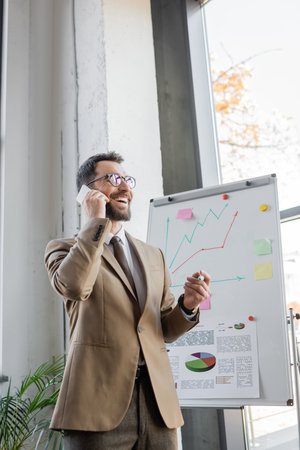 joyful bearded manager in beige blazer, tie and eyeglasses standing with marker and talking on mobile phone near flip chart with business analytics and graphs in officeの写真素材