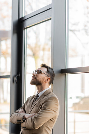 fashionable and dreamy businessman in beige blazer and stylish eyeglasses standing with folded arms near large windows and looking up in modern officeの写真素材