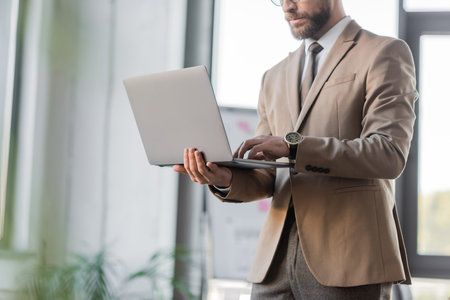 cropped view of bearded accomplished entrepreneur in beige blazer, tie and luxury wristwatch using laptop while standing in office on blurred backgroundの写真素材