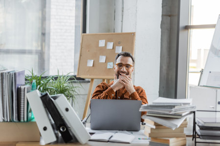 excited businessman in eyeglasses laughing and covering mouth with hands while sitting near laptop,...