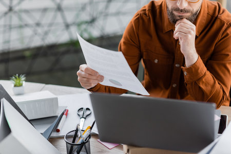 cropped view of thoughtful and busy businessman holding document near laptop, folders, scissors, marker and pen holder with pens while working at workplace in officeの写真素材