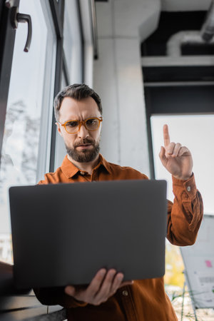 worried bearded businessman in stylish eyeglasses and shirt pointing up with finger while showing attention gesture during video conference on laptop in office, corporate lifestyleの写真素材