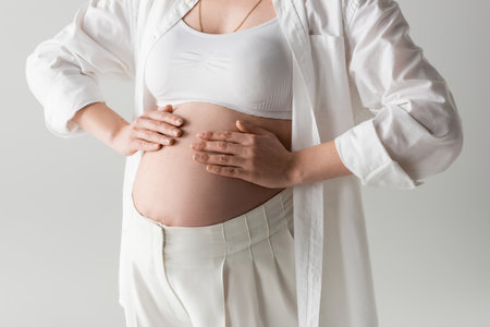 partial view of fashionable expecting mother in crop top, shirt and pants posing with hands on tummy...