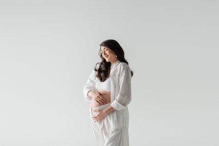 appealing and stylish pregnant model in white crop top, shirt and pants embracing tummy and smiling with closed eyes isolated on grey background, maternity fashion conceptの写真素材