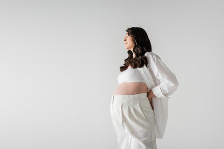 side view of smiling pregnant woman with wavy brunette hair, wearing white stylish maternity clothes, posing with hand behind back isolated on grey background, fashionable maternity conceptの写真素材