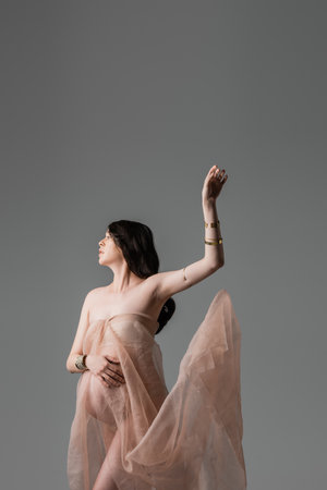 romantic pregnant woman in soft and transparent chiffon draping and golden accessories posing with raised hand isolated on grey background, maternity fashion concept, expectationの写真素材