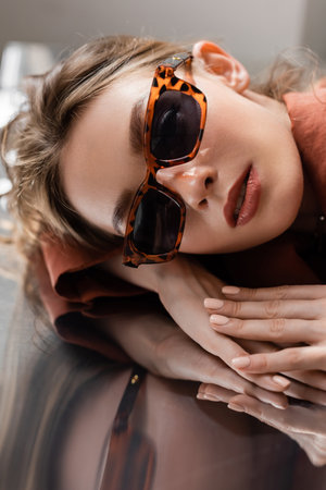 pretty young woman with brunette hair wearing trendy sunglasses and looking at camera while lying on grey mirrored background, beautiful model, reflectionの写真素材