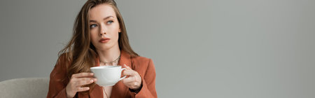Elegant and stylish brunette woman in terracotta jacket and necklaces holding cup of cappuccino and looking away while sitting on armchair isolated on grey, bannerの写真素材