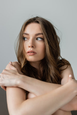 Portrait of young and pretty brunette woman with natural makeup hugging knees and looking away while sitting on grey armchair isolated on greyの写真素材