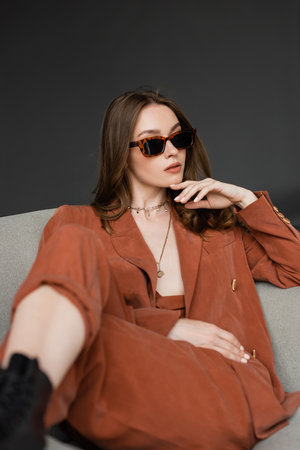 stylish young woman with long hair wearing terracotta suit with blazer and pants and posing in trendy sunglasses while sitting in comfortable armchair on grey background, fashionable model, look awayの写真素材