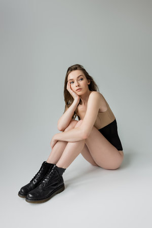 Full length of fashionable young brunette woman in black boots, corset and beige bodysuit looking away while posing and sitting on grey background, dreamy, modelの写真素材