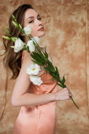 portrait of graceful, brunette and young woman in pink silk slip dress holding eustoma flowers while standing on mottled beige background, sensuality, elegance, sophistication, looking at cameraの写真素材