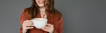 cropped view of happy young woman with brunette hair wearing brown and trendy suit with blazer and holding cup of coffee on grey background, work-life balance, bannerの写真素材