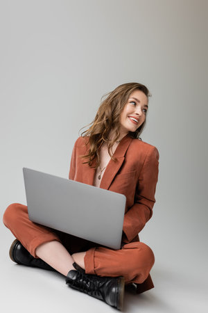cheerful young woman in golden necklace sitting with crossed legs in  trendy suit, using laptop, looking away while working remotely on grey background, freelancer, digital nomadの写真素材