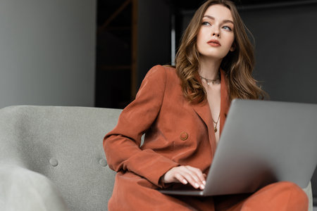 pensive young woman with long hair and necklace wearing trendy suit with blazer and pants and using laptop while sitting in comfortable armchair on grey background, freelancer, remote workの写真素材