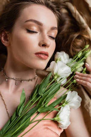 top view of attractive young woman in pink silk slip dress lying with closed eyes on linen fabric and holding white flowers on beige background, sensuality, elegance, eustoma, portraitの写真素材
