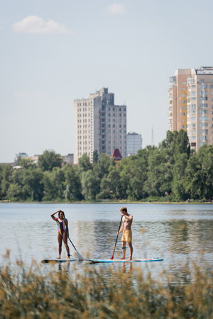 sportive interracial couple in swimwear sailing on sup boards with paddles while spending time on lake with scenic cityscape and plants on blurred foregroundの写真素材