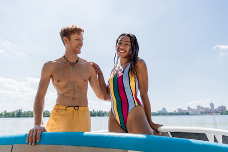 cheerful and enchanting african american woman in colorful swimsuit looking at camera near young and redhead man and sup boards under blue sky outdoorsの写真素材