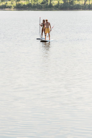 active multiethnic couple in colorful swimwear sailing on sup boards with paddles on calm river...