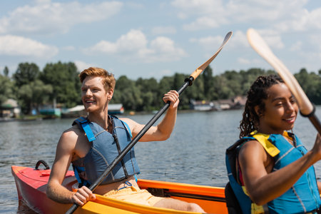 excited redhead man in life vest looking away while sailing in sportive kayak with pretty african american woman during recreation leisure on lake in summerの写真素材